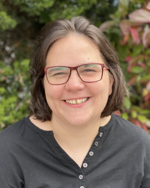 A smiling woman with shoulder-length brown hair and red-framed glasses. She is wearing a black shirt with buttons on the neckline and is standing in front of green and red foliage.
